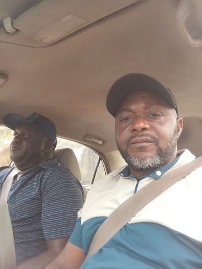 This image depicts two adult males inside a vehicle, likely in Peyer, Nigeria. The individual in the foreground, seated in the front passenger seat, is an adult male with a dark complexion, a full beard with grey streaking, and a black baseball cap. He wears a two-tone polo shirt, light beige over dark teal, and a tan seatbelt across his chest. He faces the camera directly with a neutral expression. Behind him, partially visible in the driver's seat, is another adult male with a dark complexion, wearing a dark blue baseball cap with a light-colored emblem and a dark blue horizontally striped polo shirt. He also wears a tan seatbelt and appears to be looking towards the left, not at the camera. The vehicle's interior features a beige headliner and sun visor. Blurred green foliage is visible through the rear window, indicating an outdoor setting during daylight.