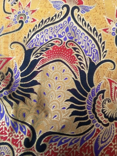 This close-up depicts a richly colored batik fabric. The dominant hues are gold, red, and indigo. Ornate patterns featuring stylized floral and foliate motifs are visible. Black outlines define the intricate shapes and swirling forms. The overall impression is one of traditional craftsmanship and vibrant, detailed design. The fabric has a slightly textured appearance.