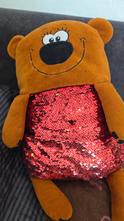 A plush toy is lying on a surface with mixed textures. The toy has a brown, soft, plush head with cartoon-like features: stitched-on eyes with eyelashes and a large black nose with a smiling mouth. The head has rounded ears. The body is covered with shiny red sequins, giving it a shimmering texture. The limbs of the toy are also made of the brown plush material. The background consists of a gray, possibly velvet-like surface and a striped, dark surface underneath the toy. The lighting suggests an indoor environment, and the focus is primarily on the plush toy. The location, Sannikovo, Russia, is not directly discernible from the visual information.