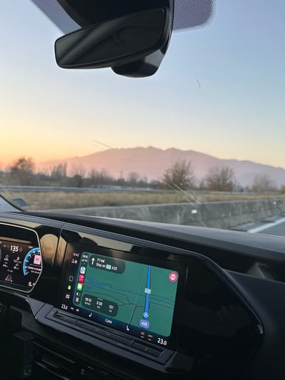 The image presents a forward-facing view from inside a moving vehicle on a highway, identified as the A25 in the vicinity of Sidirokastro, Greece. The exterior scene depicts a clear sky transitioning from pale blue overhead to warm orange and pink tones near the horizon, indicative of either sunset or sunrise. A distant, hazy mountain range forms a prominent silhouette against this colorful sky. The highway itself features multiple lanes separated by a concrete barrier, with sparse, dry vegetation lining the roadside.
Inside the vehicle, a modern dashboard is visible. The digital instrument cluster on the left displays a speed of 135 (presumably km/h). A central infotainment screen shows a navigation map with the vehicle positioned on the A25 highway, displaying a current time of 16:42, a remaining distance of 11 miles, an estimated arrival time of 18:10, and approximately 2 hours and 121 minutes of travel time. A 100 km/h speed limit sign is visible on the map. Climate control settings indicate 23.0 degrees Celsius. A rearview mirror assembly is affixed to the top of the windshield, which exhibits minor streaks and a small impact point or crack in the lower-central area. No individuals are visible within the frame.