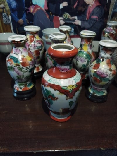 The image displays eight decorative, miniature ceramic vases arranged on a dark, polished wooden surface. The central vase features a white body with red, green, and yellow floral patterns, distinguished by a red rim and base. Seven other vases surround it, primarily white with multi-colored floral motifs in pink, orange, green, yellow, and black outlines; these typically have white rims and rest on black, tiered bases. In the blurred background, several white bowls are visible. Behind these objects, a large, out-of-focus image depicts multiple individuals in formal attire, possibly academic gowns or traditional garments, suggesting a ceremonial context. The scene is indoors, located in Steenbergen, Netherlands. No individuals are present in the foreground interacting with the vases.