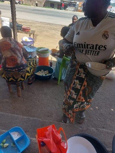 The image depicts an outdoor food vendor setting in Kimamba, Tanzania, adjacent to a paved road. In the immediate foreground, concrete steps are visible, alongside a blue segmented tray containing lime wedges and a white substance, an orange plastic basket holding red fabric, and a white paper plate resting on a dark surface.
In the midground, a woman stands, oriented towards the left, wearing a white Real Madrid "Emirates FLY BETTER" soccer jersey and a patterned sarong (khanga) in black, orange, and white. She holds a white bowl in her left hand. To her left, a person is seated on a small wooden stool, facing away from the camera, wearing a floral patterned top and a yellow and black patterned skirt. This individual is positioned near several silver pots and bowls, and a large orange insulated container. A blue basin filled with small, round, white food items (possibly dough balls) rests on the ground next to the container. A smaller individual or child is also visible, seated, holding an orange bowl and spoon.
In the background, a paved road with faint markings is present. Across the road, basic buildings and a person in a red shirt are visible. Another woman's face is partially discernible behind the standing woman, looking left. The ground is dirt, indicating an informal outdoor establishment.