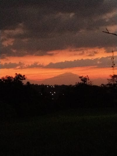 An image captured at dusk in Menjing, Indonesia, depicts a silhouetted landscape. The sky is a gradient of dark gray clouds transitioning to a vibrant orange horizon. In the distance, a mountain range is faintly visible against the fiery sky. The foreground is dominated by the dark silhouettes of trees and vegetation, with small, scattered lights suggesting human habitation or infrastructure. A tall, lattice-like structure, possibly a telecommunications tower, stands to the right of the frame. The lower portion of the image is entirely black, obscuring any details in the immediate foreground.