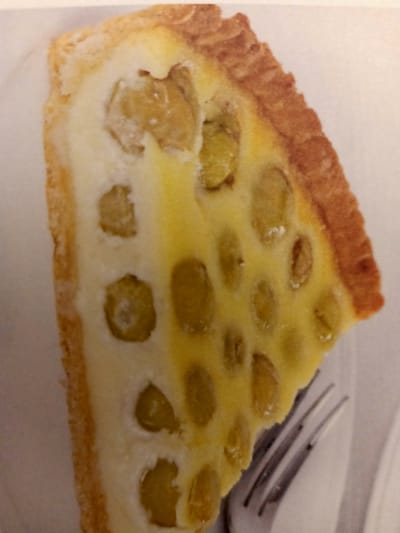 A close-up, overhead shot captures a single slice of a tart with a flaky, golden-brown crust. The filling is a pale yellow custard dotted with numerous small, round, greenish-yellow pieces, likely fruit. The tart is placed on a white surface, and a portion of a silver fork is visible in the bottom right corner. The setting appears to be a domestic environment within Rumaithiya, Kuwait.