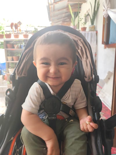 A baby is sitting in a stroller, smiling. The baby is wearing a white and striped t-shirt and green pants. The baby is holding a small piece of food in their right hand. The stroller is dark gray with a tan-colored lining. In the background, there are shelves with potted plants and what appears to be an outdoor patio area. The lighting suggests it is daytime. The overall mood is cheerful and happy, conveyed by the baby's wide smile. The scene appears to be in a home setting, likely in Panjakent, Tajikistan, given the context.