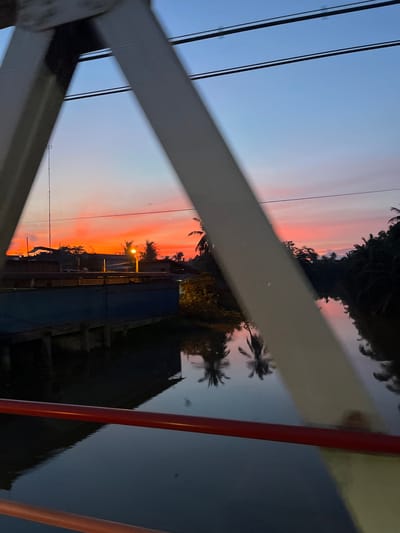 This is a photograph taken from a bridge overlooking a river at sunset. The sky displays vibrant shades of orange and pink, typical of a tropical sunset. On the left side of the image, there are buildings and structures, including what appears to be a water tower or antenna. Palm trees and dense foliage line the right bank of the river, and their reflections are visible in the water. The river itself is dark and calm, with subtle ripples. The foreground of the image is dominated by the metal structure of the bridge, including a red railing. The scene appears to be located in a tropical or subtropical region, likely a town or village situated along a river. Given the details, this could be Keudesampoiniet, Indonesia, a location known for its riverine landscapes and tropical climate. There are no people visible in the image.