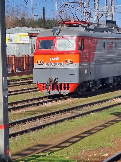 A close-up, angled shot depicts the front of a grey and red electric locomotive numbered VL10-1029. The locomotive is situated on a railway track, with greenery visible along the track's edge. Electrical power lines are overhead. A building with signs is partially visible in the background. The general context suggests the city of Fryazevo, Russia.