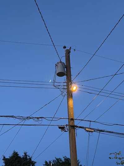 This media file captures an overhead view of a telephone pole against a clear blue sky, suggesting daytime. The pole is laden with a complex network of wires, indicative of a utility infrastructure. A prominent feature is a streetlamp mounted on the pole, illuminated and casting a warm, orange glow. Below the lamp, a grey cylindrical object, likely a transformer, is attached to the pole. Several other electrical components, including junction boxes and insulators, are visible, contributing to the dense arrangement of wires.

The presence of trees on the right side of the frame, with their dark green foliage contrasting against the sky, provides a hint of a natural setting, possibly a residential or suburban area. The sheer volume of wires and the style of the pole and equipment suggest an urban or semi-urban environment, consistent with the location context of Edmonton, Canada. The lighting from the streetlamp implies it might be dusk or early evening, despite the overall bright blue of the sky. No people are visible in the frame, and the focus is entirely on the man-made infrastructure against the natural backdrop. There is no visible text in the image. The scene is static, focusing on the static elements of the power lines and equipment.