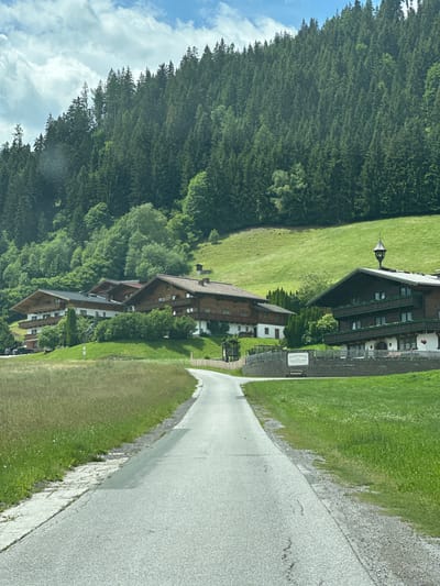 This media file captures a scenic view of Reitdorf, Austria, likely during daytime. The image is oriented vertically, with the sky and a dense forest on the left and a paved road and a row of traditional Austrian houses on the right.
The subjects are the charming wooden houses, typical of alpine architecture, with their characteristic dark wood facades, pitched roofs, and multiple windows. They are nestled on a grassy slope, with a forest of tall, dark green coniferous trees bordering the property and the edge of the sky. The sky is a bright blue, with scattered white clouds.
The scene depicts a peaceful, idyllic setting, possibly a residential area or a small resort. The road appears to be a quiet country lane, and the houses suggest a tranquil lifestyle. There's no specific activity or event taking place; the image seems to be a snapshot of the natural beauty and architectural style of the location.
Notable details include the lush green grass surrounding the houses and along the road, suggesting a well-maintained environment. The lighting indicates it's a clear day. A small, dark-colored object resembling a lamppost or a small decorative structure is visible on the grassy area near the first house.
There is some visible text on a white signpost near the entrance to one of the properties. The text is in German and appears to read "Unterbauergarten" followed by smaller text that is difficult to decipher. This sign likely indicates the name of the property or a business. The overall impression is one of serenity and natural beauty characteristic of the Austrian Alps.
