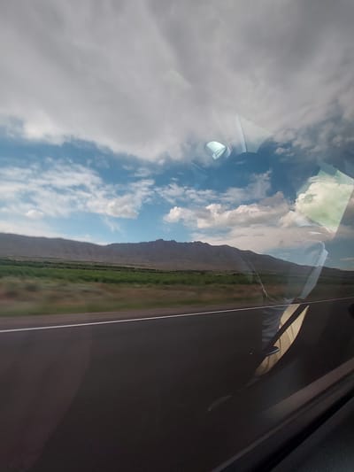 This media file captures a view from inside a vehicle, looking out through a window at a scenic landscape. The image is oriented vertically but depicts a horizontal landscape.

The upper portion of the frame is dominated by a bright blue sky with numerous scattered white clouds, some appearing large and puffy. Below the sky, a distant mountain range stretches across the middle of the image. At the base of the mountains, a strip of vibrant green vegetation, likely fields or trees, is visible, with a faint linear feature possibly indicating a road or path running alongside.

Prominent reflections on the car window overlay the exterior scene. These reflections include elements from the vehicle's interior, such as a portion of the dashboard or a speaker housing in the upper left, and the distinctive shape of a side mirror housing. Further reflections suggest the presence of a person's arm or clothing within the vehicle, particularly in the central and right parts of the image. The dark frame of the car door or window is visible along the right edge.

The scene depicts a clear, bright day, indicating sunny weather with some cloud cover. There is no visible text in the image. The landscape, characterized by mountains and green fields, suggests a rural or scenic setting, consistent with the provided location context of Paruyr Sevak, Armenia. The overall impression is that of a journey or travel through a picturesque area.