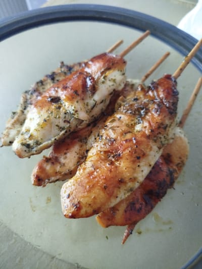 The image shows a close-up, overhead view of several chicken tenders on wooden skewers arranged on a light-colored, circular plate. The chicken tenders appear grilled, with visible char marks and browning on the surface. They are seasoned with herbs, possibly parsley and oregano, indicated by specks of green and black throughout the meat. The plate has a dark rim. The background is out of focus but suggests an indoor setting. No people are visible in the image. The scene is static and does not depict any action. The lighting is bright, creating highlights on the cooked chicken. The location is not discernable from the visual information provided in the image.