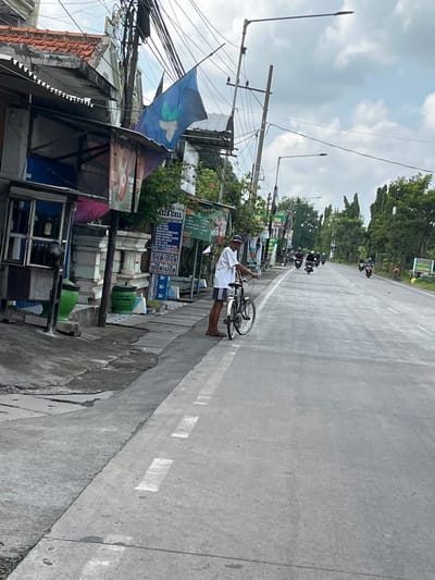The image depicts a street scene in Kepadangan, East Java. On the left side of the road, there are several buildings with awnings and signs. A man in a white t-shirt and dark shorts is standing next to a bicycle on the sidewalk. He is facing the road. In the background, there is a road with several motorcycles and riders moving in the same direction. The sky is cloudy. The environment appears to be a commercial or residential area with overhead wires and streetlights.