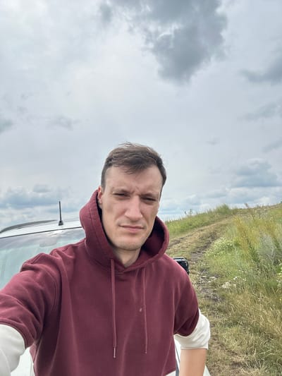 A young adult male with short brown hair and a serious expression is captured in a selfie outdoors. He is wearing a maroon hooded sweatshirt with white cuffs visible at the sleeves. The man is standing next to a white vehicle, with part of its roof and windshield visible. The background features a dirt road winding through a grassy, hilly landscape under a cloudy sky. The scene appears to be in a rural or suburban area.