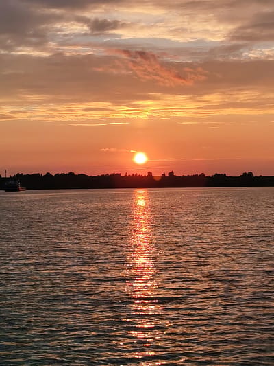 The image depicts a sunset over a body of water, likely a river or lake, with a silhouetted treeline in the background. The sun, a bright orb, is positioned just above the horizon, casting a warm, orange glow across the sky and reflecting a shimmering path of light on the water's surface. The sky is filled with a mix of light and dark clouds, tinged with shades of orange and pink. In the distance, along the treeline, a boat can be seen to the left, its form partially obscured by the twilight. The water itself appears rippled, creating a textured surface that catches the sunlight. The scene evokes a sense of calm and natural beauty. The location is identified as the city of Selivanovo, Russia.