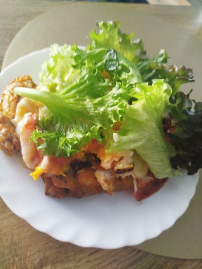 A close-up shot shows a white plate filled with food, positioned on a wooden surface. The plate is round and has a wavy edge. The food appears to be a type of layered dish, possibly a sandwich or open-faced creation. It includes a base layer that looks like fried or breaded material, topped with pieces of shrimp, and a generous portion of fresh green lettuce leaves. Some red and yellow ingredients are also visible within the dish, possibly tomatoes and a sauce. The overall impression is of a light meal. The lighting suggests it might be daytime, possibly indoors given the controlled setting of a plate on a table. There are no people visible, and no text is present in the image. The scene is simple and focused solely on the presented food item.
