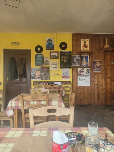 This image depicts an indoor dining area or tavern located in Κωστήτσι, Greece. The space features walls painted bright yellow on the left and center, transitioning to vertical wood paneling on the right. The ceiling is white, from which a black geometric light fixture hangs. The floor is dark, appearing to be tiled or paved with stone.
A brown wooden door is situated on the left, with an "ΕΞΟΔΟΣ" (EXIT) sign mounted above it. Two dark coats hang on hooks on this door, and a key, possibly accompanied by an umbrella, dangles from the door handle. On the right, a darker wooden door is visible, partially obscured, with a white "W" sign affixed, indicative of a restroom.
The walls are extensively decorated with various posters, primarily in Greek, advertising plays, films, and products. Notable posters include "Ο ΣΦΑΚΙΑΝΟΣ Χ.Σ.ΛΑΜΣ," "Μέρες μαΝάργιας," "Ο ΓΛΑΡΟΣ," "ZOO," "ΚΟΛΥΠΟΣ" (featuring a woman in a red dress), "ΔΗΠΕΘΕ ΙΩΑΝΝΙΝΩΝ "ΟΙ ΕΛΛΗΝΕΣ"," "Ο ΕΜΠΟΡΟΣ ΤΗΣ ΒΕΝΕΤΙΑΣ," a vintage "KODAK FILM" advertisement, and a "ΚΑΡΕΛΙΑ" cigarette advertisement showing a woman smoking. Two black vinyl records are mounted on the yellow wall. A small wooden shelf on the wood-paneled wall holds a light-colored, elongated gourd. A light switch and electrical outlet are visible on the yellow wall section adjacent to the wood paneling.
The room contains multiple light-colored wooden chairs with woven straw seats. Tables are covered with plaid tablecloths featuring red, yellow, and white patterns. In the immediate foreground, a table is partially visible, displaying a red container (possibly a napkin dispenser or ashtray), a small clear glass bottle, two clear glasses containing water, and a plate holding a white, crumbled food item. A fork is placed near the plate, and an ashtray with a cigarette butt is visible behind the bottle. No individuals are present in the scene, and no actions or interactions are depicted. The arrangement of items on the foreground table suggests recent occupancy or preparation for use.
