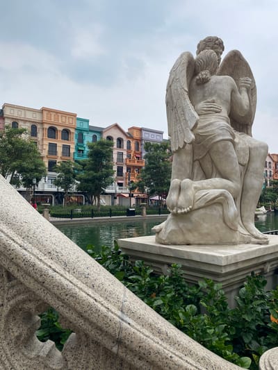 The image depicts a bright, overcast day in Nghĩa Trụ Commune, Vietnam. In the foreground, a large, light-colored stone statue of two figures embracing, one with feathered wings, is positioned on a marble pedestal. Lush green bushes surround the base of the statue. Behind the statue, a body of water reflects the sky and surrounding structures. Further in the background, a row of vibrantly colored buildings, in shades of peach, orange, teal, and purple, with architectural details reminiscent of European townhouses, line the waterfront. Trees with full green canopies are interspersed between the buildings and the water. A stone railing with ornate carvings is visible in the lower left quadrant of the image, partially obscuring the view of the buildings and water. The overall scene suggests a well-maintained public space, possibly a park or entertainment district.