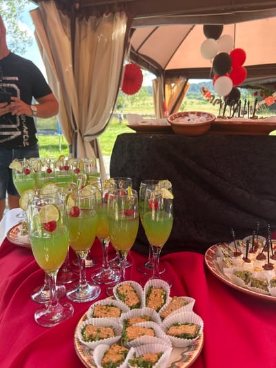 This image captures a vibrant outdoor event, likely a party or celebration, set in a garden or yard. The primary subjects are a table laden with refreshments and a group of people in the background.

On the table, several glasses of a bright green beverage are meticulously arranged. Each glass is garnished with a slice of lime and a cherry, suggesting a festive cocktail. Alongside the drinks, there are plates of appetizers. One plate holds small, rectangular sandwiches or canapés, each wrapped in white paper liners and topped with a garnish of green herbs. Another plate displays various small bites, including some that appear to be dried meats and others with a green paste, all artfully arranged on skewers. A bowl of ice is also visible.

In the background, a person wearing a black t-shirt with visible text is partially visible, adding a human element to the scene. Other people are also present, though less distinctly. The setting features a tent or canopy, adorned with beige fabric, creating a shaded area. Red paper fans are used as decorations. A bunch of balloons in red, white, and black colors are tied to a stand, further enhancing the celebratory atmosphere. The overall impression is one of a well-catered outdoor gathering. The lighting suggests it is daytime, likely with pleasant weather. The location context of Gorni Okol, Bulgaria, places the event in a specific geographical setting.
