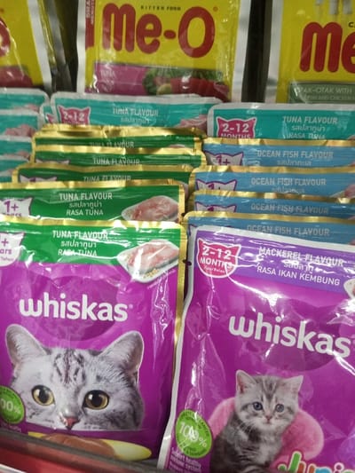 The image depicts an indoor retail display of cat and kitten food products within a store in Naung Bo Oe, Myanmar. In the foreground, two large purple Whiskas brand pouches are prominently featured: one displaying a grey tabby cat's head and the other a grey tabby kitten, both advertising "Complete & Balanced 100%." Behind them, multiple smaller individual wet food pouches are vertically stacked. On the left, green-topped pouches are labeled "TUNA FLAVOUR" (1+ Years), and on the right, blue-topped pouches are labeled "OCEAN FISH FLAVOUR" (2-12 Months), with a partially visible purple "MACKEREL FLAVOUR" pouch. In the background, yellow bags of Me-O kitten food, including "TUNA" and "OTAK-OTAK WITH FISH AND CHICKEN" varieties, are arranged on shelves. The setting is brightly lit, and no people are visible in the frame.