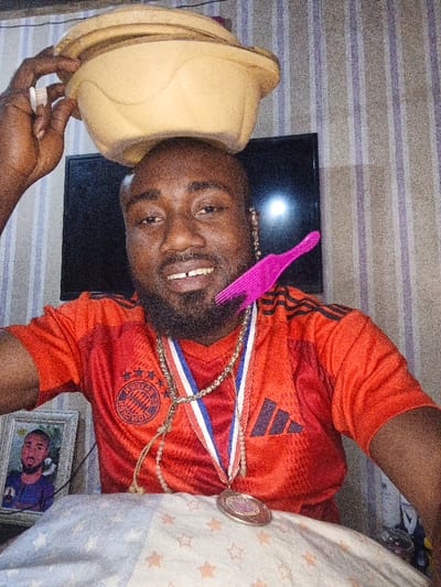 A man balances stacked bowls on his head. He wears an orange sports jersey, with several necklaces including a medal, and has a pink comb tucked by his ear. A framed portrait sits to his left. A black television is behind him, and the background is vertically striped wallpaper.