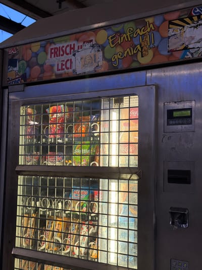 The image shows a close-up view of a vending machine that appears to be located in Witten, Germany. The machine is filled with various snacks and drinks, including bags of Skittles, Haribo Tropifrutti, and Coca-Cola bottles. The top of the machine features a colorful advertisement with German text that reads "Frisch & Leck" and "Einfach genial!". The machine itself is made of metal and has a glass display window showing the products inside. A digital display on the right side of the machine shows "EUR 0,00" and some German text. The overall environment suggests an outdoor setting, possibly a street or a public area, given the lighting and the slightly weathered appearance of the machine and its surroundings. There are no people visible in the image.