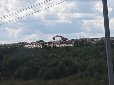 An excavator is visible on a large mound of debris, possibly a landfill, under a cloudy sky. The excavator is yellow and appears to be in operation. The debris mound is covered with light-colored material, possibly trash or soil. In the foreground, there is dense green vegetation, including trees and bushes, and a dirt path. A concrete pole is on the right side of the image, and power lines are visible in the upper part of the image. The location is Ivanovka (Verevskoe S/P), Russia.