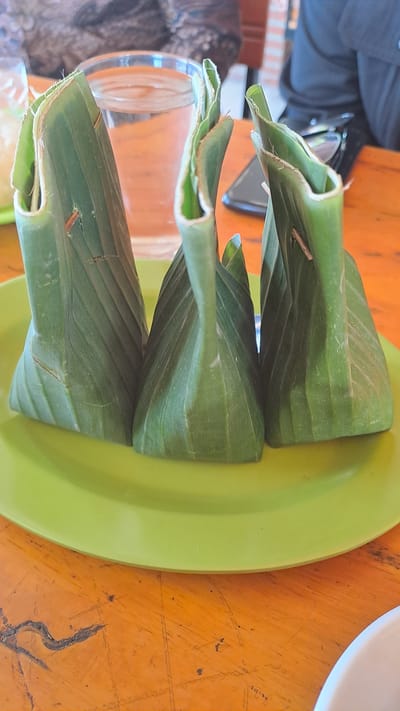 Three rice parcels, wrapped in banana leaves and shaped into triangular packets, sit on a lime-green plate. The plate rests on a worn, orange wooden table, the surface of which is marked with scratches and dark stains. In the background, a glass of clear liquid with ice cubes is visible, next to a portion of a person wearing a black shirt. Another person, partially obscured, wears a patterned brown and black top. The scene appears to be set in an indoor location, possibly a restaurant or dining area, in Julikeude Dua, Indonesia. A small, light brown insect is perched on the left-most banana leaf packet.