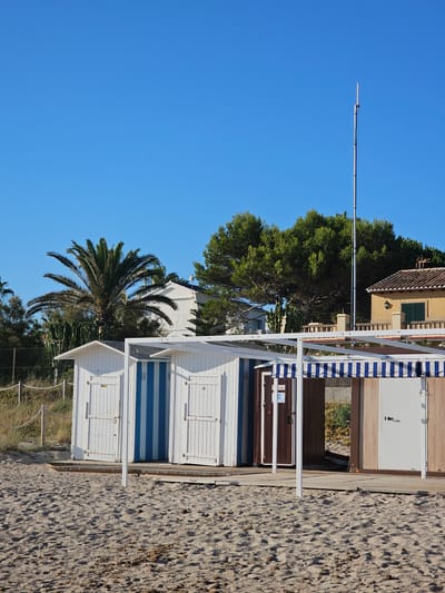 The image depicts a row of beach huts on a sandy beach under a clear blue sky. The beach huts are constructed of wood and painted white, with some featuring blue vertical stripes. One hut on the right has a brown wooden facade and a white door, covered by a blue and white striped awning. A tall antenna stands prominently in the background, behind a dense green tree line. In the middle ground, a white building with a palm tree in front is visible, along with other vegetation. The foreground is dominated by the sandy texture of the beach, with small pebbles and debris scattered across it. The scene is set in Deveses, Spain. There are no people visible in the image.