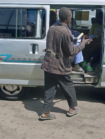 In Communauté rurale de Payar, Senegal, an outdoor daytime scene depicts a male adult standing beside an open white passenger van on an asphalt surface. The man, seen from a three-quarter rear view, wears a worn brown and tan plaid jacket with a strap-like feature on the lower back, dark trousers, and gray slide sandals. He holds a light-colored, elongated object, possibly a folded paper or small book, in his left hand while extending his right hand towards the interior of the van.
The white van's right-side sliding door is fully open. A passenger inside the van, wearing a light blue garment and bright green slide sandals, is visible, with their arm extended towards the man outside, suggesting an exchange. Further inside the vehicle, another individual is partially visible. The van's window immediately behind the driver's side features extensive spiderweb-patterned cracking, indicative of impact damage, though the glass remains in place. A light blue stripe runs along the lower body of the van. Bright sunlight casts distinct shadows on the ground.