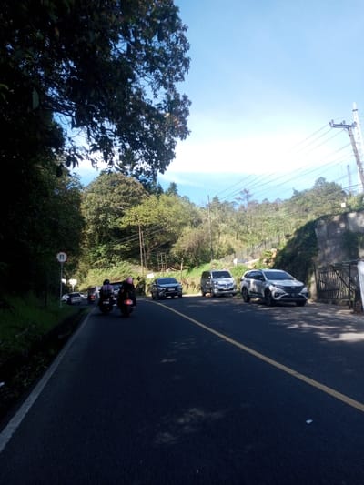 This image depicts a two-lane asphalt road traversing a mountainous or hilly area in Ka Thaung (Upper), Myanmar, under a clear blue sky. The road features a solid white line on its left shoulder and a solid yellow line delineating the center. Two individuals on motorcycles are traveling away from the viewer in the left lane; one rider wears a pink helmet, the other a dark helmet, and both motorcycles appear to carry rear cargo. A queue of additional vehicles is visible further ahead in the same lane. On the right side of the road, three passenger vehicles are parked parallel to the road: a dark-colored SUV, a silver minivan, and a light-colored SUV. Dense green foliage and trees line both sides of the road, extending up the steep hillsides. A circular white sign with a red border and the number '11' is visible on the left side of the road. Utility poles and power lines run along the right side of the road, adjacent to a concrete retaining wall on the ascending terrain. A narrow drainage ditch is present along the left edge of the road. The scene is illuminated by direct sunlight.