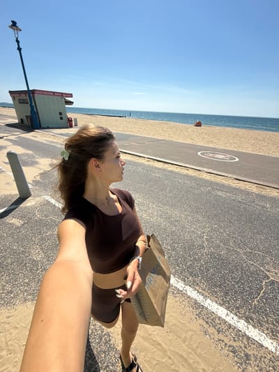 A medium-shot selfie shows a young woman with brown hair, wearing a brown crop top and shorts, standing on a sandy path next to a paved walkway. She is holding a brown paper shopping bag from Primark in her right hand, and her left arm is extended towards the camera. The woman is looking to her right, towards the sea. Behind her, a beach hut with a sign that reads "Hot & cold drinks, snacks, ice creams" is visible, along with a tall lamppost. The sea is calm and blue, meeting a sandy beach. A paved path runs parallel to the beach, and a circular marking that appears to be a helipad is visible on the path. The sky is clear and blue. The overall scene depicts a sunny day at the beach in Bournemouth, United Kingdom.