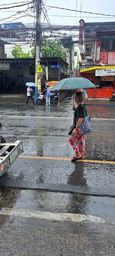 The image captures a street scene in Mandaluyong, Philippines, during a rainy day. The foreground is dominated by a wet asphalt road with a distinct orange line marking a lane. Raindrops create ripples on the puddles formed on the road.

In the middle ground, an elderly woman is seen walking, holding a light blue umbrella overhead to shield herself from the rain. She is dressed in a black shirt and a vibrant pink and floral-patterned skirt, carrying a patterned blue bag over her shoulder. Beside her, further down the street, another person walks with a smaller, light blue umbrella. A motorcycle is parked nearby, and another individual in a light blue shirt and dark pants is standing beside it, holding an umbrella.

The background reveals a bustling street with buildings characterized by red and yellow facades. A dense network of electrical wires hangs precariously above the street, a common sight in urban Philippines. Signs are visible, including a yellow sign with two figures that appears to be a pedestrian crossing or warning sign. Another sign, partially obscured, reads "CARWASH OPEN 24 HOURS." The overall atmosphere suggests a typical urban environment dealing with inclement weather, with the rain adding a layer of subdued activity. The time of day appears to be daytime, possibly late morning or afternoon, given the ambient light.
