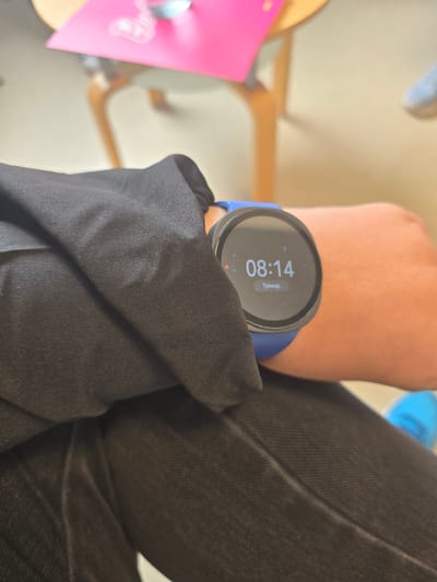 A person's arm is visible with a smartwatch on the wrist. The watch face displays the time as 08:14. The arm is partly covered by a black fabric, likely clothing. A leg is also visible, wearing dark-colored jeans. In the blurred background, there is a pink object on a wooden chair. This could be located in Oberasbach, Germany, but without more information, this is not conclusive.