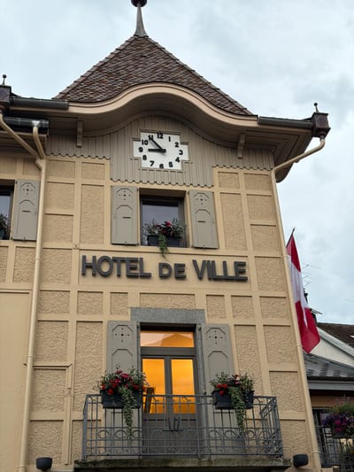 The image shows the facade of a building identified by the signage "HOTEL DE VILLE," indicating it is the town hall. The building has a distinctive architectural style with a textured beige exterior and darker, grid-like decorative elements. Above the "HOTEL DE VILLE" sign, there is a clock face with Roman numerals. There are windows with shutters and planters filled with red flowers on a balcony. A Swiss flag is visible on the right side of the building, attached to a downspout. The sky is overcast. The scene is located in Saint-Gervais-les-Bains, France.