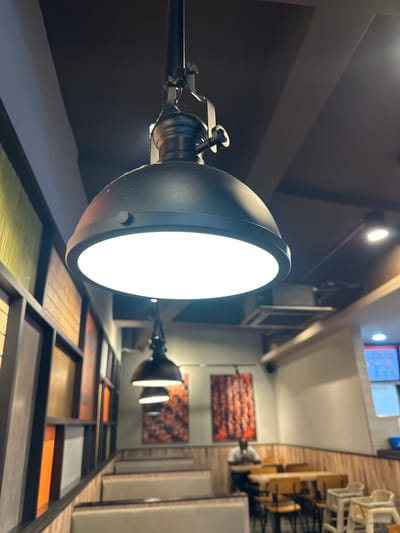 This image depicts the interior of a dining establishment located within the city of Subang Jaya City Council, Malaysia. The perspective is low-angle, looking upwards towards the ceiling and across the seating area.
In the immediate foreground, a prominent black, bell-shaped pendant light fixture hangs from a dark-colored ceiling, illuminated brightly from its base with a stark white light. Fine dust particles are visible on the upper surface of the fixture. The suspension mechanism, dark and metallic with an industrial design, connects the fixture to the ceiling.
To the left, a decorative wall feature runs horizontally, composed of a dark-framed grid containing multiple rectangular panels in varying muted colors, including green, yellow, orange, brown, and white. Below this feature, a row of booth-style seating with light beige upholstery and wooden bases is arranged.
Further into the room, two additional pendant light fixtures, similar in style but smaller and further recessed, are suspended from the ceiling. On the far wall to the right, two vertically oriented framed artworks are displayed, featuring abstract patterns predominantly in shades of red and orange. Below the artworks, a dining area is furnished with multiple light-colored wooden tables and chairs. A single individual is seated alone at one of these tables, their head bowed towards the tabletop. Adjacent to the dining tables, several light-colored high chairs are stacked.
On the far right, a service counter or bar area is visible, featuring a blue container and illuminated digital screens displaying colorful graphics. The ceiling throughout the establishment is dark, likely black or dark grey, with some lighter-colored structural beams or panels visible. Additional recessed lighting fixtures and an air conditioning vent are present on the ceiling. The lower portion of the walls beneath the artworks and service area is paneled with light-colored wood, while the upper walls are painted a neutral light grey or off-white.