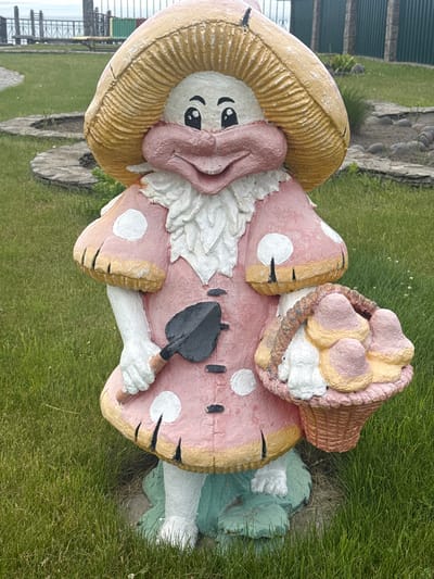 An outdoor, eye-level, full-shot photograph captures a whimsical mushroom-shaped garden gnome statue situated in a grassy area, possibly a park or yard in Kuril'sk, Russia. The gnome, predominantly pink and yellow, wears a large, textured mushroom cap as a hat and a pink dress adorned with white polka dots and yellow trim. It has a smiling face with big black eyes and a pink nose. The gnome holds a small black trowel in its right hand and a woven basket filled with pastel-colored mushroom-shaped objects in its left. The statue stands on a green base, partially obscured by grass. The background includes a stone pathway, more green grass, and a metal fence with a glimpse of water and distant structures.