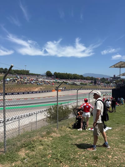 The image captures a sunny day at the Circuit de Barcelona-Catalunya, located near Granollers, Spain. In the foreground, a chain-link fence separates the viewer from a section of the race track, characterized by its gray asphalt and vibrant green runoff area. Beyond the track, a large crowd of spectators is visible, many seated on a grassy hillside dotted with colorful umbrellas. The background features trees, distant mountains, and a clear blue sky with wispy white clouds. Several individuals are positioned in the foreground, some standing and observing the track, while others are tending to a stroller. The overall scene depicts a busy event at a prominent motorsport venue.