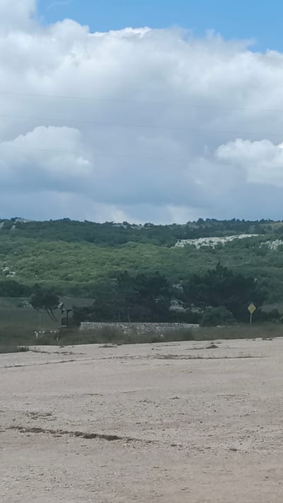 The image depicts a wide, dirt or gravel surface in the foreground, appearing to be a road or parking area. In the middle ground, there is a low, rough-stone wall running horizontally, behind which a small, wooden, covered structure, possibly a shelter or gazebo, is visible. A few dark green trees are situated around this structure. Further back, the landscape rises into rolling hills covered in dense, light-green vegetation, suggesting a forested or scrub-covered area. In the distance, the terrain is more rugged with lighter patches of rock or exposed earth. The sky above is a mix of blue and white clouds. There are no people or animals visible in the frame. The scene appears to be in a natural or semi-natural outdoor environment.