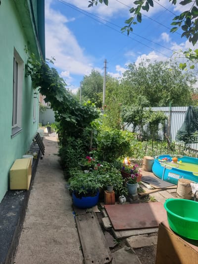The image shows a view of a backyard in Saran, Kazakhstan. The foreground features a concrete walkway with a light green building on the left, possibly a house. A yellow plastic storage container is placed on a dark, raised base against the building's wall. Various potted plants, including flowers in shades of pink, red, and purple, are arranged along the walkway. To the right, a bright blue inflatable swimming pool is visible, filled with water and colorful plastic balls, suggesting it's a child's play area. A white fence with vertical corrugated metal panels borders the backyard. Lush green trees and foliage are abundant, indicating a summer season. The sky is blue with scattered white clouds. Electrical wires are visible overhead, strung between utility poles. The overall scene depicts a residential backyard setting, possibly during daylight hours.