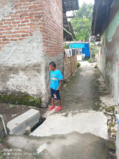 A young boy stands in an alleyway wearing a blue shirt, shorts, and red sandals. He is positioned near a corner where a brick and concrete wall meets a concrete path. The alley narrows into the distance with a bamboo fence on one side and a weathered wall on the other. In the background, a blue structure is visible. The ground is damp with patches of moss and a concrete drainage system in the foreground.