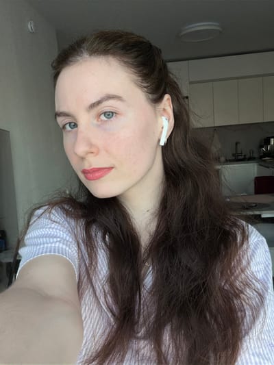 The scene is an indoor portrait of a young adult female within a residence in Ковалёво, Russia. The female subject presents with fair skin, blue eyes, and long, dark brown, wavy hair, partially pulled back from her face. She is wearing pink lipstick and a light-toned, striped collared shirt. A white wireless earbud is inserted into her right ear. The subject directly faces the camera, indicating a selfie. The background consists of a modern kitchen, featuring white upper cabinetry, a stainless steel sink with a faucet, and visible countertop appliances. A circular, flush-mount ceiling light fixture is present. Lighting indicates a combination of natural ambient light from an unseen left source and interior artificial illumination.
