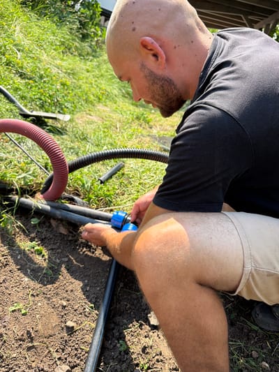 In Bašť, Czechia, a bald man with a beard is kneeling in a grassy, dirt area, focused on connecting a blue plastic fitting to black pipes. Two flexible hoses, one red and one black, are visible in the foreground, lying near the pipes. The man is wearing a black polo shirt and light-colored shorts. The scene appears to be outdoors, with sunlight casting shadows on the ground.