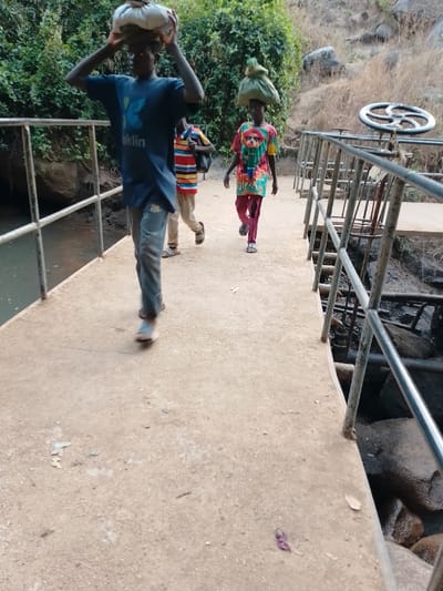 The image captures an outdoor scene featuring three young individuals walking across a concrete bridge over a body of water. The bridge has metal railings on both sides.
In the foreground, a young person, possibly a male, is walking towards the viewer, slightly blurred by motion. They are wearing a dark blue t-shirt with a light blue logo and the word "aklin" printed below it, light-colored jeans that appear somewhat distressed or worn, and light-colored slip-on shoes or sandals. This individual is balancing a large, light-colored sack or bundle on their head, holding it steady with both hands.
Behind them and slightly to the right, another young person, also walking towards the viewer, is clearly visible. They are wearing a vibrant, colorful t-shirt with a busy pattern that includes shades of yellow, green, red, and black, featuring what appears to be an animal or character design on the front. They are also wearing reddish-pink trousers with white stripes on the side and white slip-on sandals. This person is expertly balancing a green, irregularly shaped bundle or sack on their head without using their hands.
Further back and to the left of the second person, a third individual is partially visible, wearing a striped t-shirt (red, orange, blue, white stripes) and light-colored trousers or shorts, with what seems to be a dark backpack. They are also wearing black and white sandals.
On the left side of the bridge, a dark body of water or stream can be seen below the railing. The bank beyond the water is covered in dense green foliage and trees. On the right side of the bridge, below the railing, there are rocks and what appears to be dark, possibly muddy ground. A large, circular metal wheel, resembling a valve or gate control, is prominently positioned on a metal structure attached to the right side of the bridge. The background on the right features a dry, rocky hillside with sparse vegetation.
The concrete surface of the bridge appears dusty and somewhat worn, with small bits of debris like leaves and a crumpled purple item scattered on it. The scene is brightly lit, indicating daytime. The individuals appear to be engaged in carrying goods, a common activity in many regions.