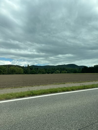 The image presents a serene landscape captured from a roadside perspective. In the foreground, a gray asphalt road with a distinct white line marking its edge occupies the right side of the frame. Adjacent to the road is a narrow strip of green grass, which transitions into a wider area of tilled, dark brown earth, indicative of a field.

Beyond the field, a series of rolling hills or mountains covered in dense, dark green trees forms the mid-ground and background. The tree cover appears lush and unbroken along the hillside. Above the mountainous terrain, the sky is largely overcast, dominated by various formations of clouds. Some clouds are thin and streaky, while others are thicker and more diffused, with patches showing brighter light, suggesting breaks in the cloud cover or the sun's presence behind them. A small section of blue sky is faintly visible amidst the clouds.

The scene is devoid of any visible people, animals, or specific activities, portraying a tranquil, natural environment. The time of day appears to be daytime, under mostly cloudy conditions. No text is visible in the image. The setting is consistent with rural or semi-rural areas, and the provided context places this scene near Zurzach, Switzerland.