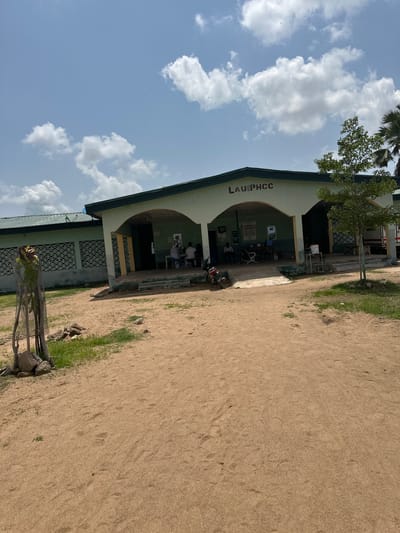 The image shows a building with the text "LAU PHCC" visible on its facade. The building appears to be a school or medical facility, with an arched entrance leading to an interior area where a few people are seated. Outside the building, there is a sandy area with some sparse grass and vegetation. The sky is blue with scattered white clouds. A tree with green foliage is growing near the building. The scene suggests a daytime setting, likely in a warm climate, given the sandy terrain and clear sky. The overall impression is one of a functional, possibly public, building in an outdoor, somewhat arid environment.