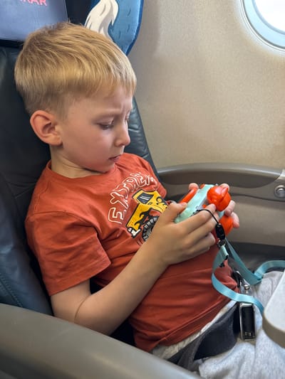 The image shows a young boy lying down on a seat, likely on an airplane, given the presence of a window and a seatbelt. He is wearing an orange t-shirt with a graphic design featuring yellow and white text and imagery. His hands are holding a small, colorful toy. The boy has blonde hair and appears to be relaxed or possibly sleeping. The background includes airplane interior elements like a seatback and a window. The lighting suggests it is daytime. The overall scene conveys a sense of travel and a child occupied with a toy. There is no text that is clearly legible in the image.