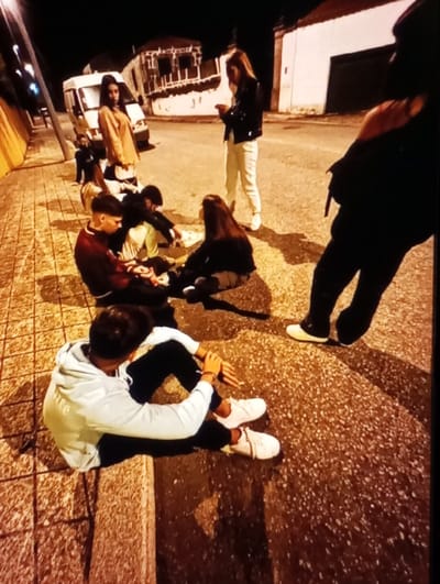 This is a nighttime image depicting a gathering of approximately eight young individuals on a street in Aktash, Uzbekistan.
The scene features a paved sidewalk on the left and an asphalt road on the right, separated by a concrete curb. Elongated shadows are cast from the left, indicating an unseen light source. In the background, a white van is parked on the road, behind which a building with a dark facade and white trim is visible. To the far right in the background, another building with a white wall and a dark garage door or entrance stands.
From left to right and foreground to background, the individuals are positioned as follows:
1. A male is seated on the sidewalk with his legs bent and arms resting on his knees. He wears a light-colored hooded sweatshirt, dark pants, and white sneakers, with his head slightly bowed.
2. Adjacent to him, another male is seated on the sidewalk, wearing a dark red long-sleeved top and dark pants, appearing to look down at his lap.
3. Behind these two, a female with long dark hair is seated on the ground, wearing a dark top and light-colored pants.
4. Next to her, another individual, likely male, is seated on the ground, wearing a light-colored top and dark pants.
5. Standing in the road, a female wearing a dark jacket, light-colored pants, and white sneakers is actively using a mobile phone.
6. To her left, partially on the road, a female is standing, dressed in a light brown long-sleeved top and a white skirt or pants.
7. Further back on the left, a partially obscured individual in dark clothing is seated or crouching on the sidewalk, behind the white van.
8. In the foreground on the right, a female, largely cropped and facing away, stands with one shoulder exposed, wearing a dark top and dark pants, with white shoes visible.
The individuals are clustered together, with some seated on the ground and others standing. No direct conversations or interactions are overtly visible; however, their proximity suggests a group gathering.