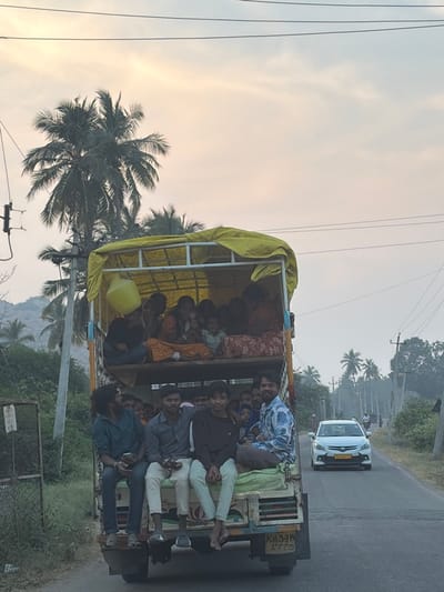 A light-duty truck, painted light blue-green with a yellow tarpaulin covering its upper structure, is observed from the rear on a paved road in Chikkarampur, India. The truck is heavily laden with approximately 20-25 individuals. The upper section, beneath the yellow tarp, contains an estimated 10-15 people, predominantly women and children, seated on what appears to be a raised platform. One person in this section holds a yellow plastic jerrycan. The lower rear platform of the truck carries approximately 8-10 young adult males, seated with their legs dangling off the back. One male on the far right is smiling, while another on the far left holds a mobile phone. The truck's license plate, "KA53M 2775," is visible at the bottom rear.

The scene is set on a two-lane asphalt road lined with dense green foliage and numerous tall palm trees. Utility poles carrying power lines are visible on the right side of the road. Distant hills are present on the left horizon. The sky is overcast with a pale, soft light, suggesting either early morning or late afternoon. A white sedan, bearing license plate "KA04MJ6399," approaches in the opposite lane.