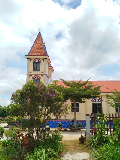 The image depicts a brightly colored church with a tall, slender bell tower, located in Xuân Sơn, Vietnam. The church has a yellow facade with a red tile roof. The bell tower is also yellow and topped with an orange pyramidal roof. It features decorative windows with stained-glass accents. In front of the church, there is a lush garden with a variety of plants, including a tree with purple flowers. A dirt path leads towards the church. The sky is partly cloudy with a mix of blue sky and white clouds. The overall impression is one of a serene and well-maintained religious building in a tropical setting.