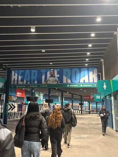 The image captures an indoor concourse or hallway within a large venue, identified as Wembley in the United Kingdom. The ceiling features dark, parallel panels with integrated lights, casting an artificial glow consistent with an evening or night setting.
Along one wall, a prominent blue banner displays the phrase "HEAR THE ROAR" in large white letters, alongside an image of a male soccer player in a white jersey. Below this, various green directional signs indicate services such as "PROGRAMMES," "MERCHANDISE," "↑ WATER POINT," "↑ INFORMATION," and "+ FIRST AID." A red sign reads "HERE TO TAP," likely for a payment or water station, while another green sign shows a female restroom symbol and the number "108," possibly indicating a gate or section.
Several people are visible, primarily from behind, suggesting movement through the concourse. In the foreground, individuals wearing dark jackets and casual clothing are walking. Further back, more people are seen near what appear to be kiosks or service counters. One person with long, wavy, reddish-brown hair is prominent among the crowd. In the distance, near the center-right, a person in a dark jacket stands, possibly a staff member, observing the flow of people.
The scene depicts attendees at a large public event, most likely a sporting event given the context of Wembley and the "HEAR THE ROAR" banner with a soccer player. The various service signs confirm the operational nature of the venue during an event.